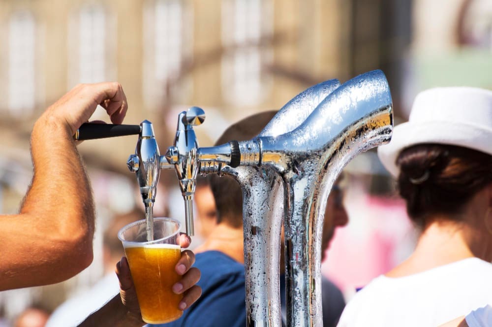A hand pulls a lever on a shiny chrome beer tap, pouring golden beer into a clear plastic cup, with blurred people and a building in the sunny background, evoking a lively outdoor event.