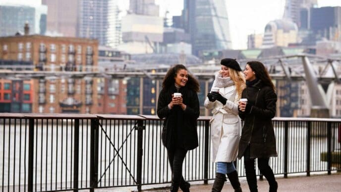 Three smiling female friends, dressed in warm winter coats, walk along a riverside promenade, holding coffee cups, with modern city buildings and a bridge in the blurred background under a grey sky.