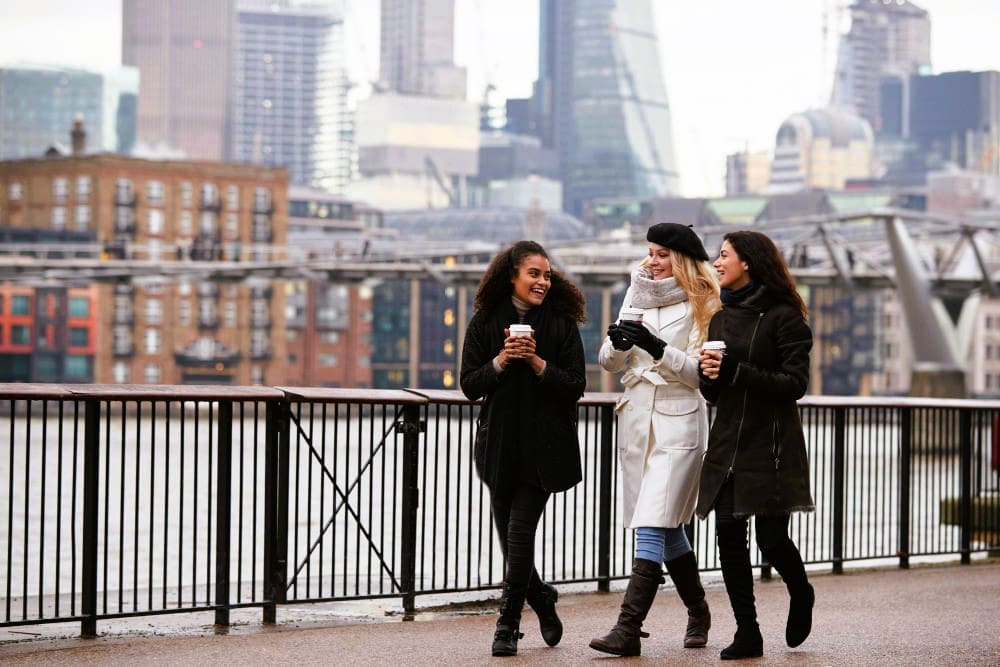 Three smiling female friends, dressed in warm winter coats, walk along a riverside promenade, holding coffee cups, with modern city buildings and a bridge in the blurred background under a grey sky.