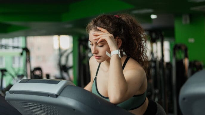 A young woman who appears to be White, with long, curly hair, leans on a treadmill console with her hand on her forehead, looking tired or frustrated in a gym.