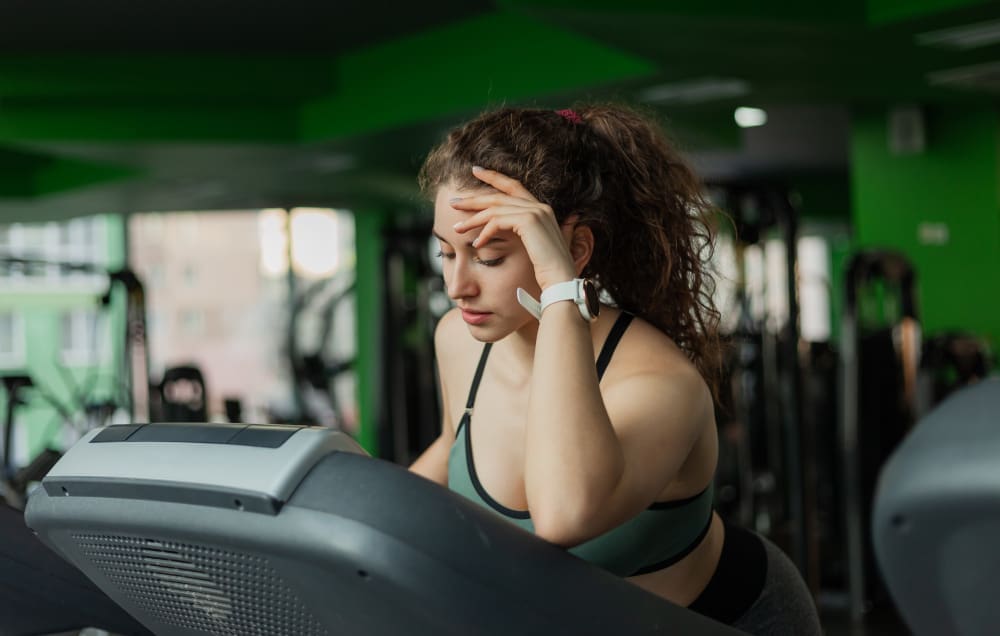 A young woman who appears to be White, with long, curly hair, leans on a treadmill console with her hand on her forehead, looking tired or frustrated in a gym.