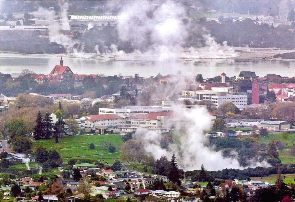 An aerial view of Rotorua, New Zealand, showing a city nestled around a lake, with numerous plumes of white steam rising from geothermal activity amidst buildings and trees, under an overcast sky.