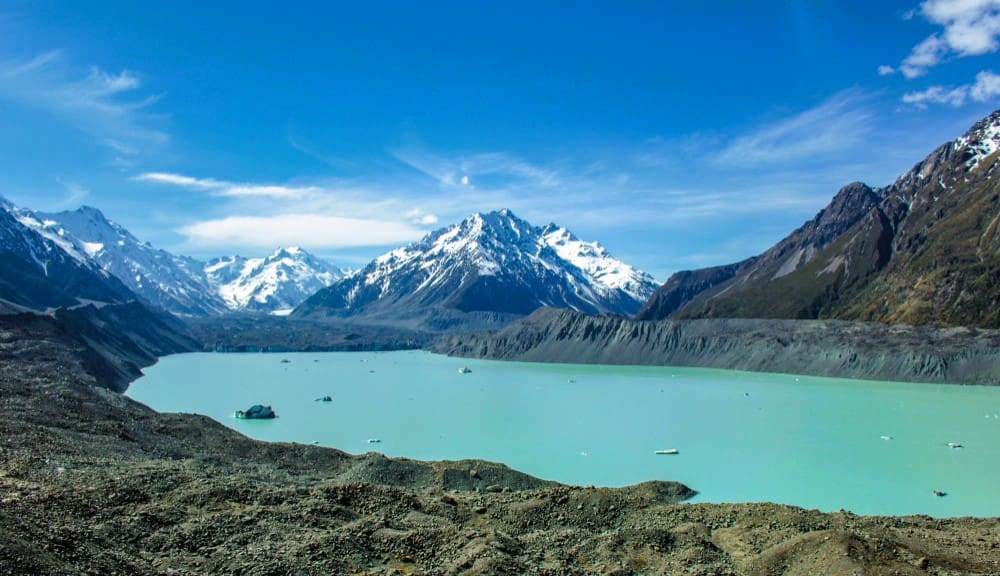 A panoramic view of a large, serene turquoise glacial lake surrounded by dark, rocky moraines and towering snow-capped mountains under a clear blue sky in Aoraki / Mount Cook National Park, New Zealand.