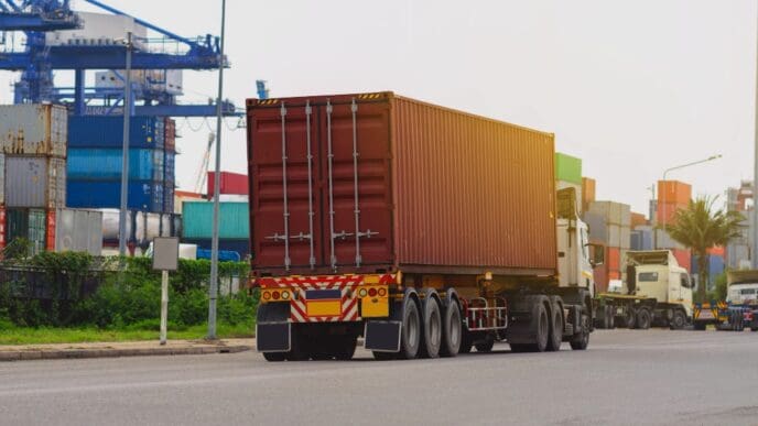The rear view of a large red shipping container on a truck driving on a road, with a bustling port in the background featuring stacked containers and large cranes under a clear sky.