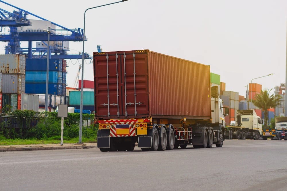 The rear view of a large red shipping container on a truck driving on a road, with a bustling port in the background featuring stacked containers and large cranes under a clear sky.