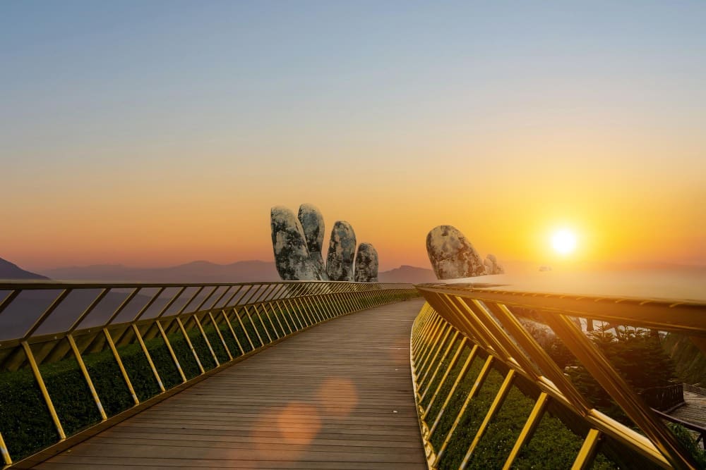 The Golden Bridge (Cau Vang) in Vietnam, held by giant stone hands, stretching into the distance at sunset with mountains in the background.