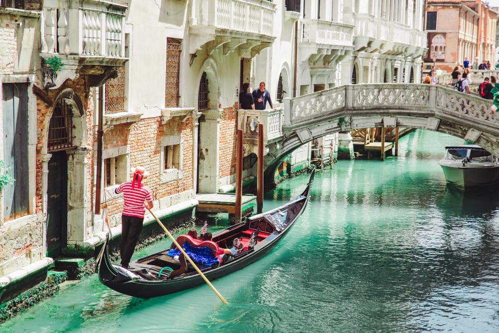 A gondolier in a striped shirt steers a black gondola on a turquoise canal in Venice, Italy, surrounded by historic buildings and a stone bridge.