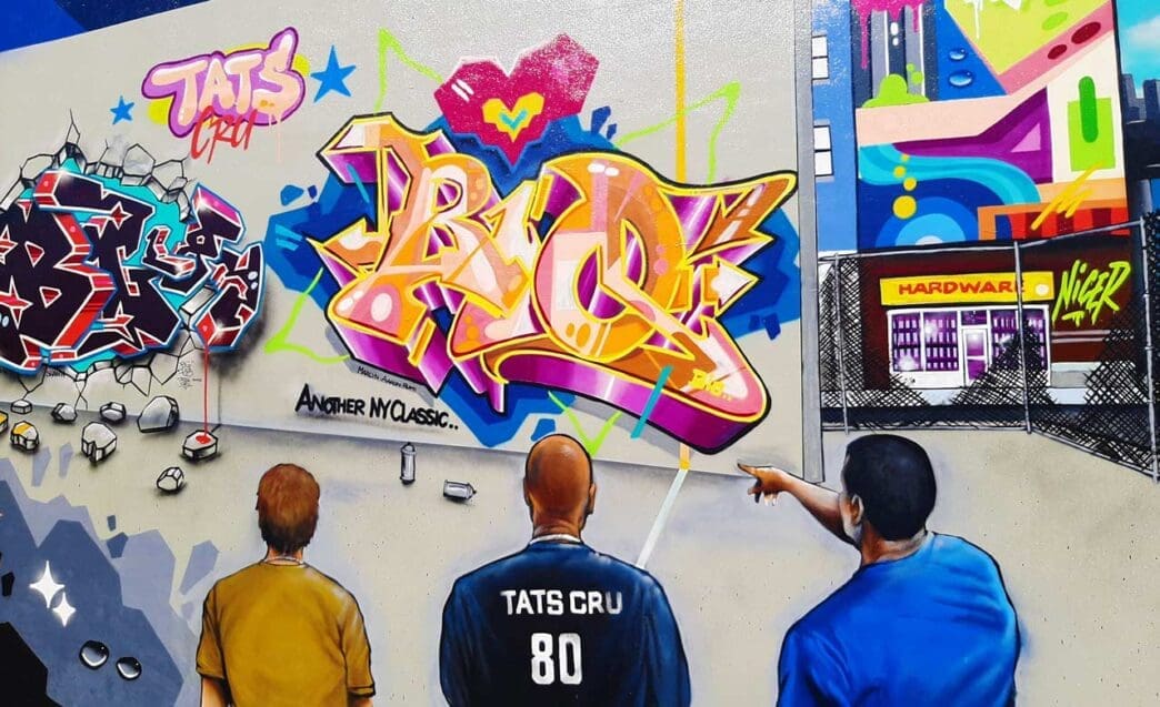 A vibrant graffiti mural in Wynwood, Miami, featuring three people looking at a large, colorful piece of "TATS CRU" graffiti lettering. The mural incorporates a broken-wall effect and a detailed cityscape with a "HARDWARE" store.