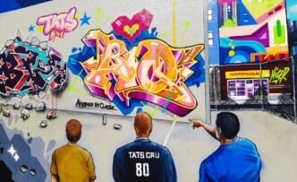 A vibrant graffiti mural in Wynwood, Miami, featuring three people looking at a large, colorful piece of "TATS CRU" graffiti lettering. The mural incorporates a broken-wall effect and a detailed cityscape with a "HARDWARE" store.