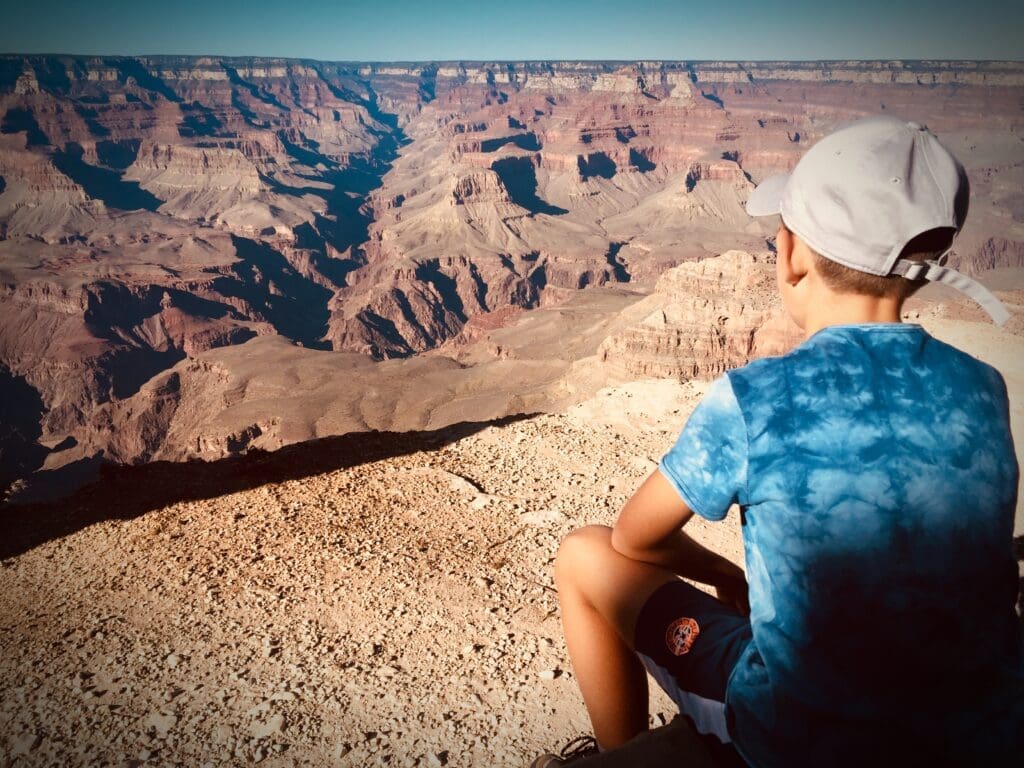 Rear view of a young boy in a cap and tie-dye shirt, sitting and gazing out at the vast Grand Canyon from its rim under a clear sky.