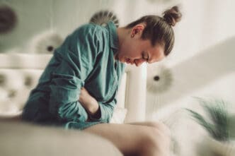 A young woman sits hunched over with her arms wrapped around her stomach, her eyes closed and face contorted in pain or discomfort, symbolizing gut issues or abdominal pain.