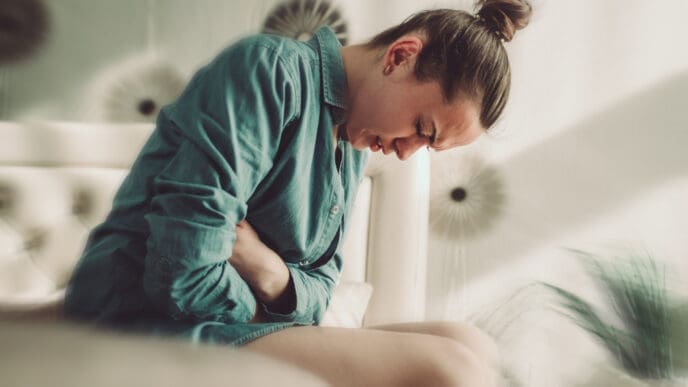 A young woman sits hunched over with her arms wrapped around her stomach, her eyes closed and face contorted in pain or discomfort, symbolizing gut issues or abdominal pain.
