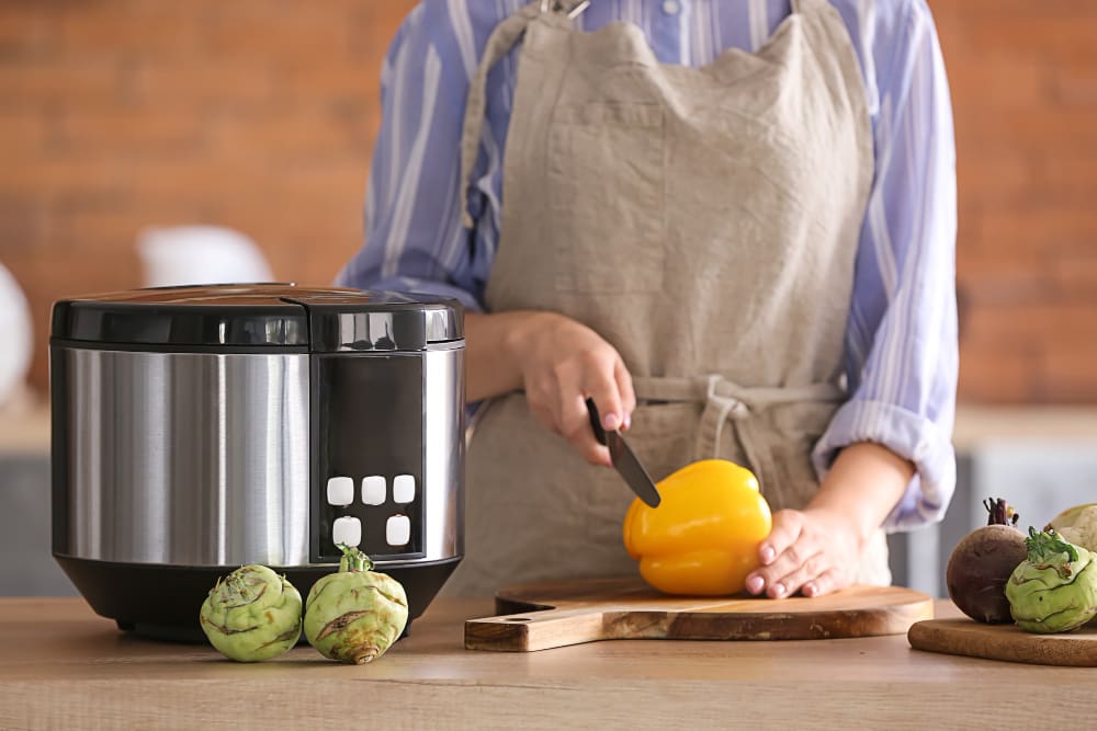 A woman in an apron cutting a yellow bell pepper on a wooden board next to a modern multi-cooker in a kitchen.