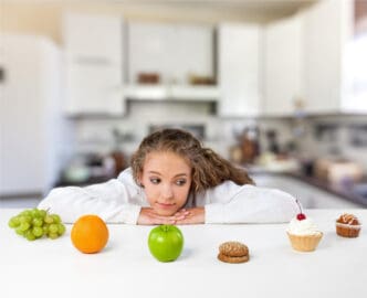 A young woman lies on a counter, resting her chin on her forearms and looking intently at a selection of healthy fruits (apple, orange, grapes) and unhealthy treats (cupcake, cookies, muffin), symbolizing food choices and healthy habits.