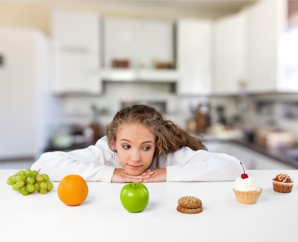 A young woman lies on a counter, resting her chin on her forearms and looking intently at a selection of healthy fruits (apple, orange, grapes) and unhealthy treats (cupcake, cookies, muffin), symbolizing food choices and healthy habits.
