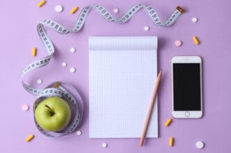 A top-down flat lay of health and wellness items on a purple background, including a blank notepad, a pencil, a green apple with a white measuring tape, a smartphone, and scattered pills and capsules.