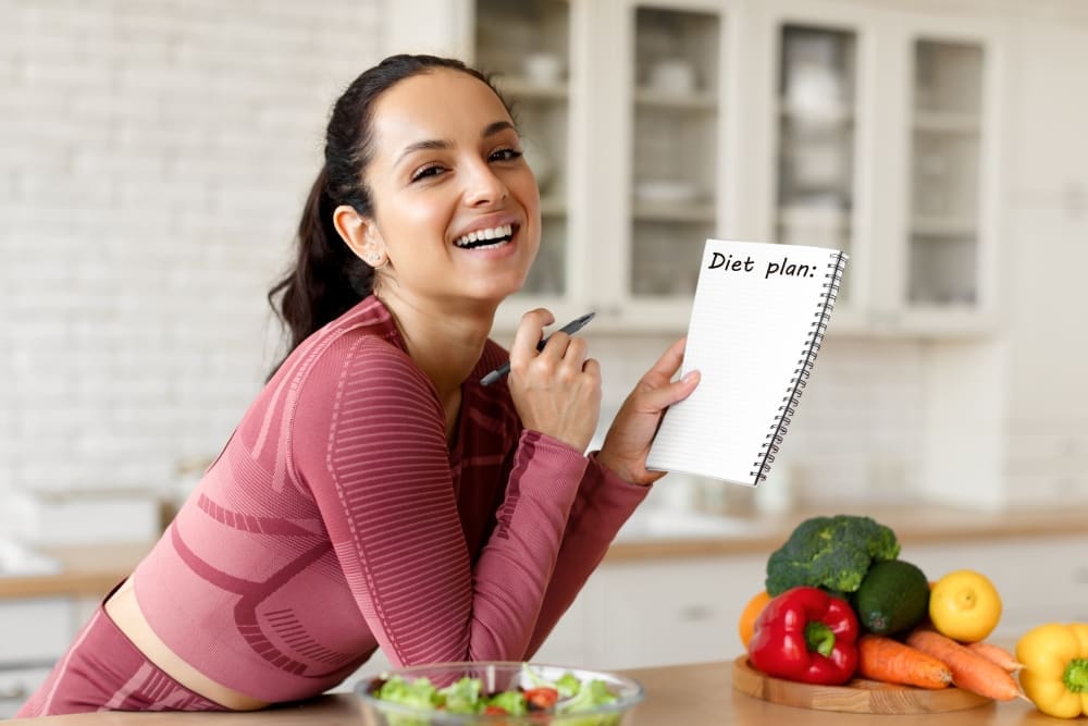 A happy young woman who appears to be Hispanic or Mediterranean, smiles while leaning on a kitchen counter, holding a notebook titled "Diet plan:" and a pen, with fresh vegetables and a salad nearby.