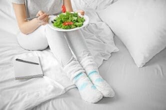 A woman sitting in bed in comfortable loungewear and fluffy socks, eating a fresh green salad from a white plate, with a notepad and pen nearby, symbolizing a healthy routine.