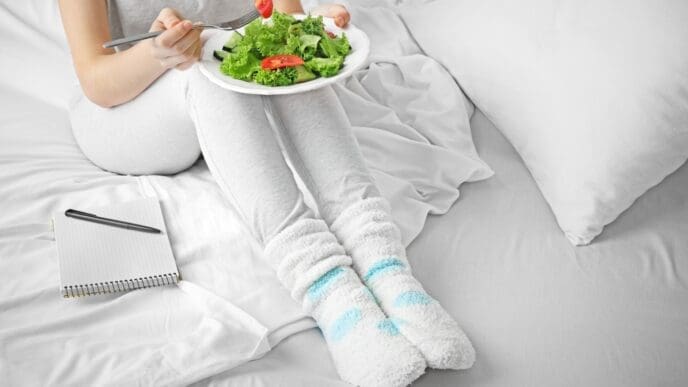 A woman sitting in bed in comfortable loungewear and fluffy socks, eating a fresh green salad from a white plate, with a notepad and pen nearby, symbolizing a healthy routine.