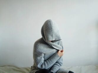 A person wearing a large, gray hooded sweater sits on a bed with their face completely hidden by the hood and their arms wrapped around their body, symbolizing withdrawal or hidden anxiety.