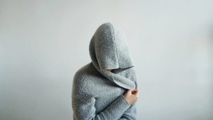 A person wearing a large, gray hooded sweater sits on a bed with their face completely hidden by the hood and their arms wrapped around their body, symbolizing withdrawal or hidden anxiety.