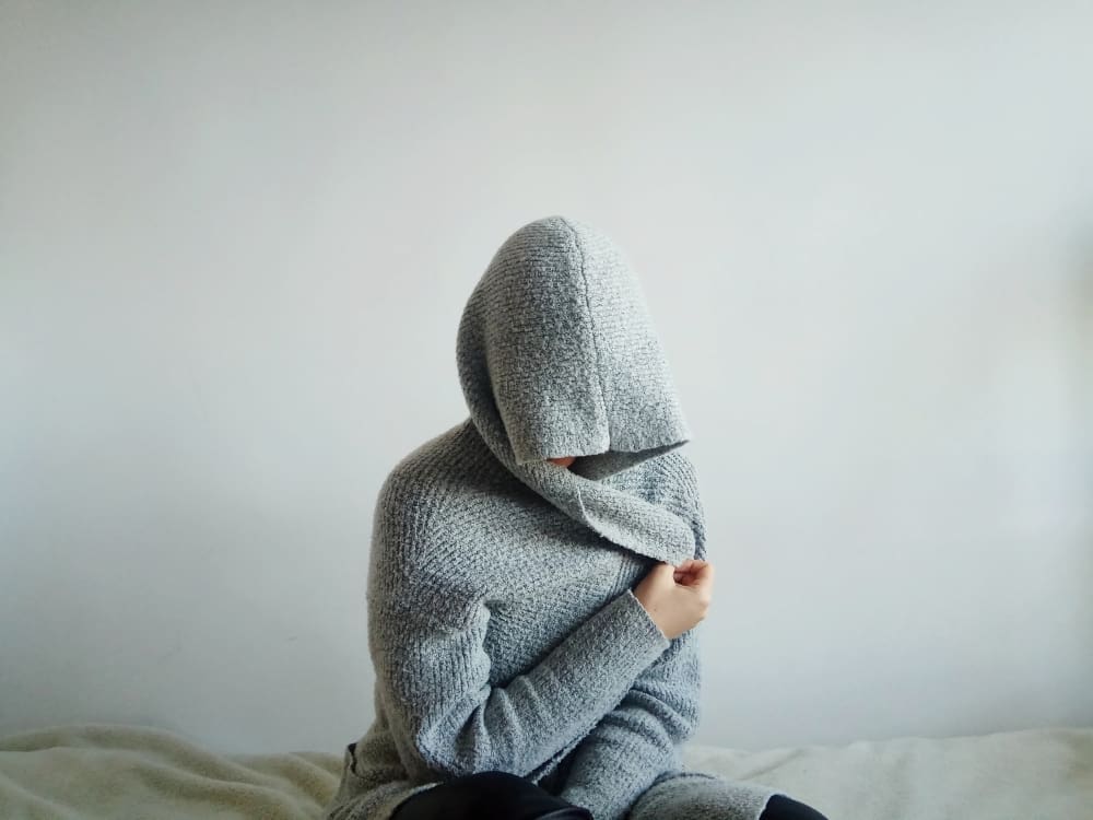 A person wearing a large, gray hooded sweater sits on a bed with their face completely hidden by the hood and their arms wrapped around their body, symbolizing withdrawal or hidden anxiety.