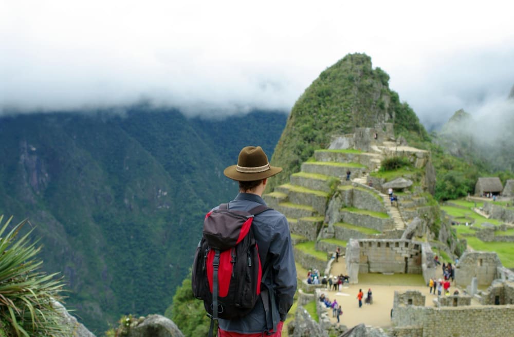 A hiker wearing a brown hat and a red and black backpack stands with their back to the camera, looking out over the ancient stone terraces and structures of Machu Picchu, with mist-shrouded green mountains in the background.