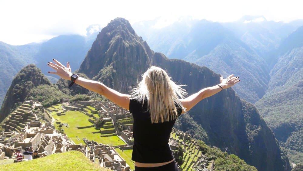 A blonde woman stands with her back to the camera, arms outstretched, overlooking the ancient Inca city of Machu Picchu and the majestic Huayna Picchu mountain, under a bright sky with distant hazy mountains.