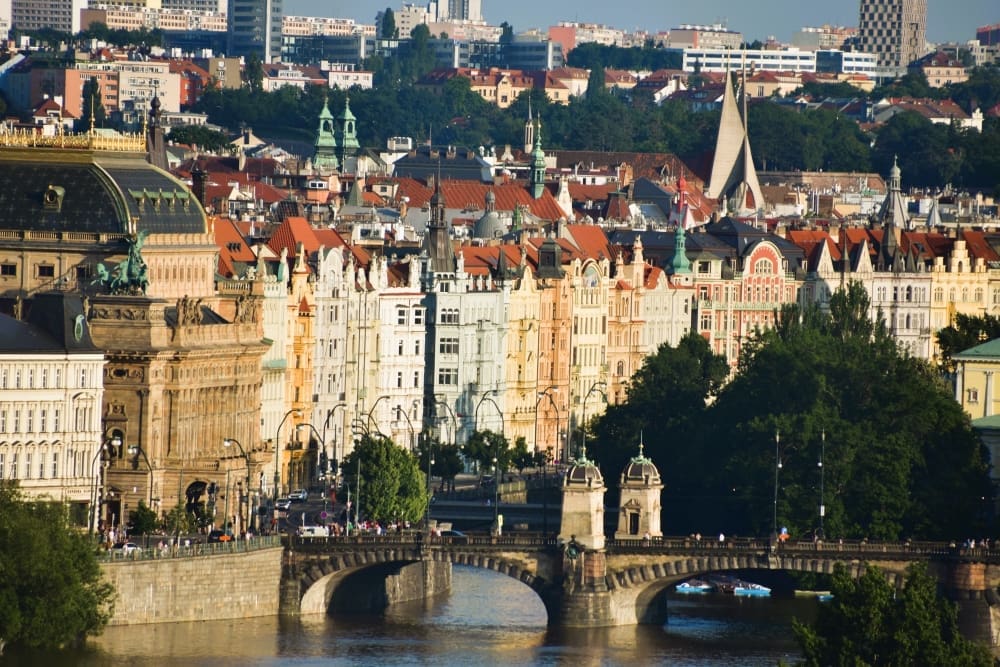 Panoramic view of Prague with historic buildings, a bridge over the Vltava River, and a modern spire in the distance.