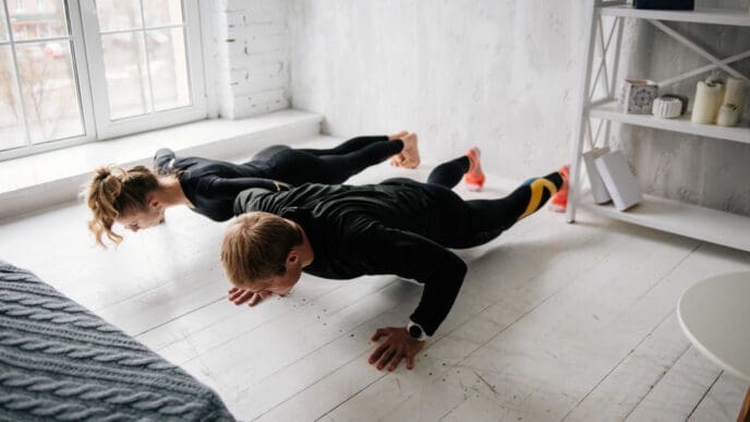 A man and a woman performing push-ups side-by-side on a light wooden floor in a bright room, demonstrating bodyweight exercise.