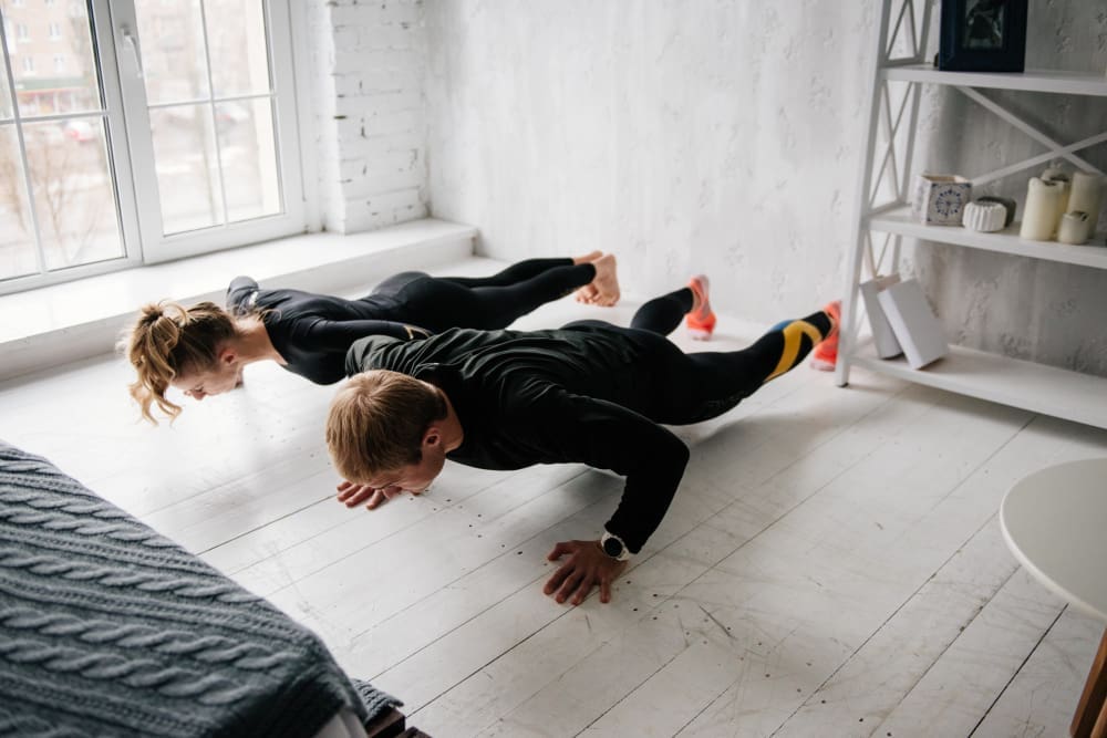 A man and a woman performing push-ups side-by-side on a light wooden floor in a bright room, demonstrating bodyweight exercise.
