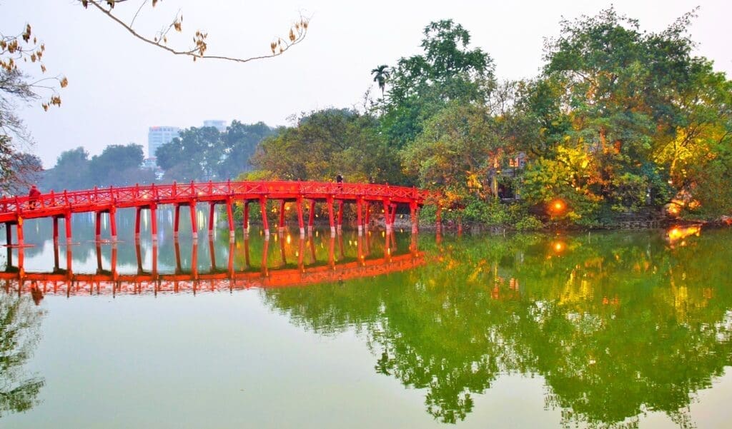 The vibrant red Huc Bridge reflecting in the calm waters of Hoan Kiem Lake, surrounded by green trees.