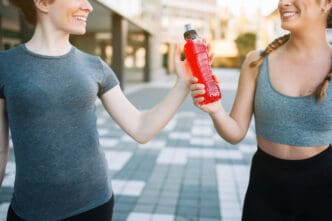 Two smiling female runners, one in a grey t-shirt and the other in a grey sports bra, share a red electrolyte drink bottle on a paved outdoor surface.