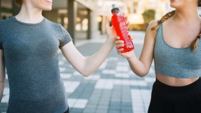 Two smiling female runners, one in a grey t-shirt and the other in a grey sports bra, share a red electrolyte drink bottle on a paved outdoor surface.