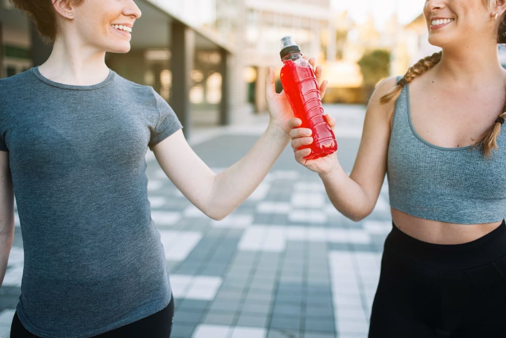 Two smiling female runners, one in a grey t-shirt and the other in a grey sports bra, share a red electrolyte drink bottle on a paved outdoor surface.