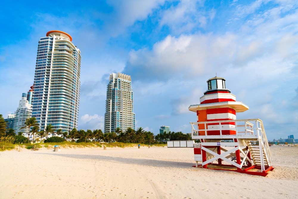 A red and white striped lifeguard tower on South Beach, Miami, with towering skyscrapers and palm trees in the background under a blue sky.