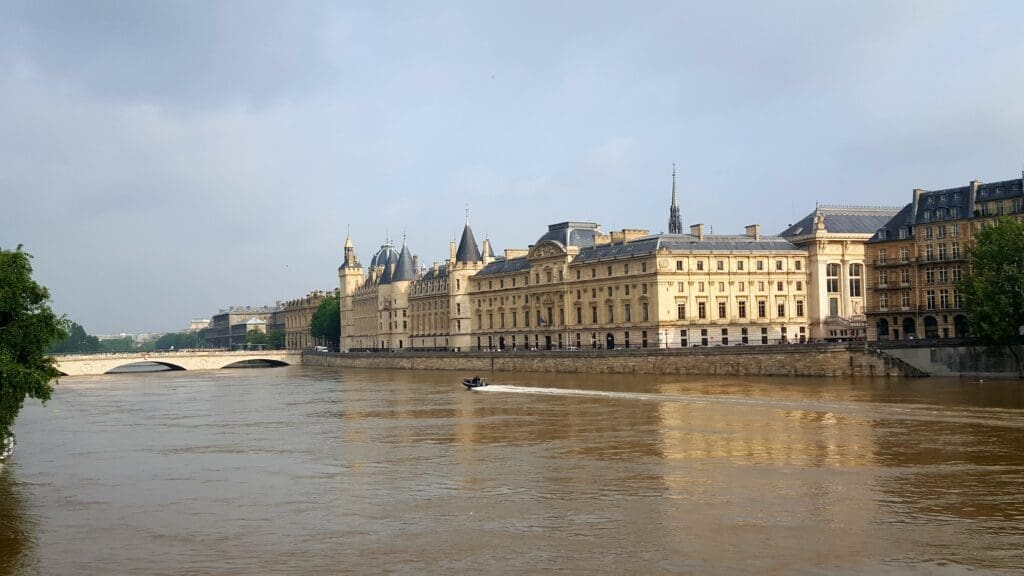 A panoramic view of historic buildings along the Seine River in Paris, including the Conciergerie and Palais de Justice on Île de la Cité, with a bridge and a small boat on the brownish water under a cloudy sky.