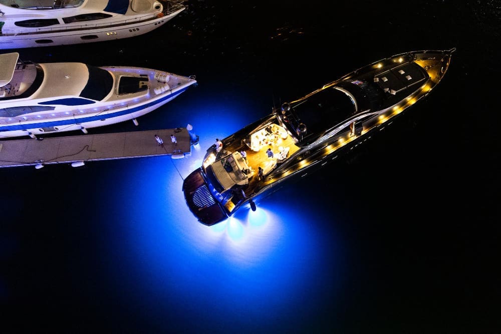 An aerial view of a luxurious yacht illuminated with bright blue underwater lights at night, docked near other yachts.