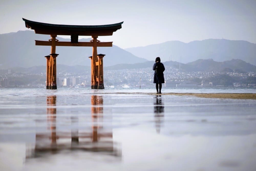 The iconic red torii gate of Itsukushima Shrine stands in the shallow waters of Miyajima Island, Japan, with a lone person looking towards it and distant mountains visible across the bay.