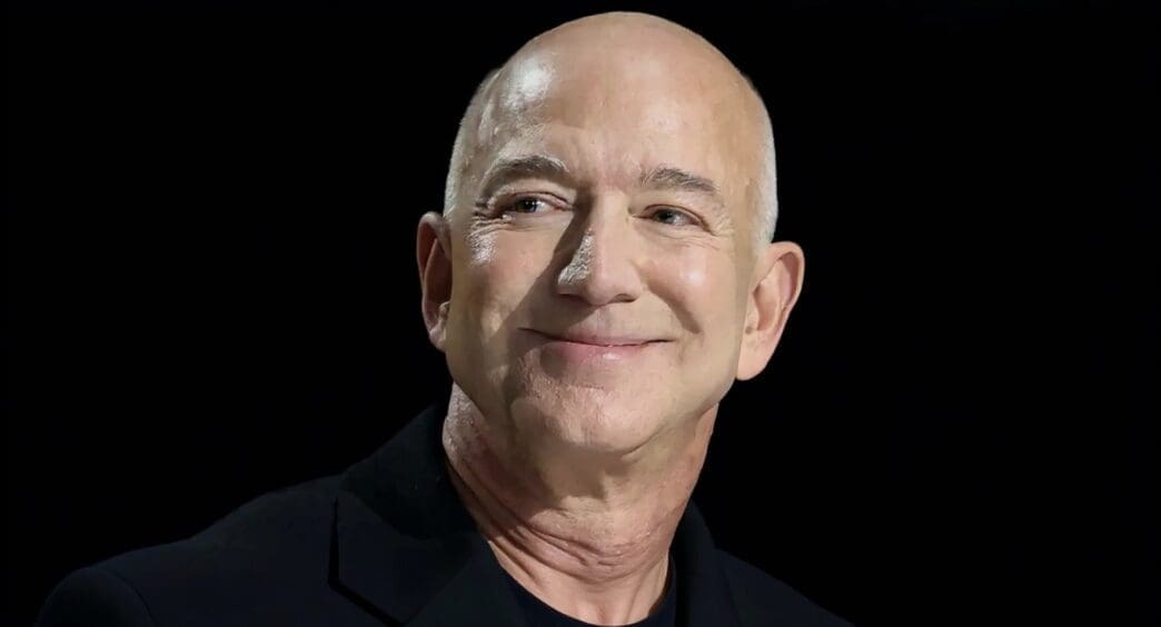 A close-up portrait of Jeff Bezos, bald and smiling, looking slightly upwards and to his left, against a dark, blurred background.
