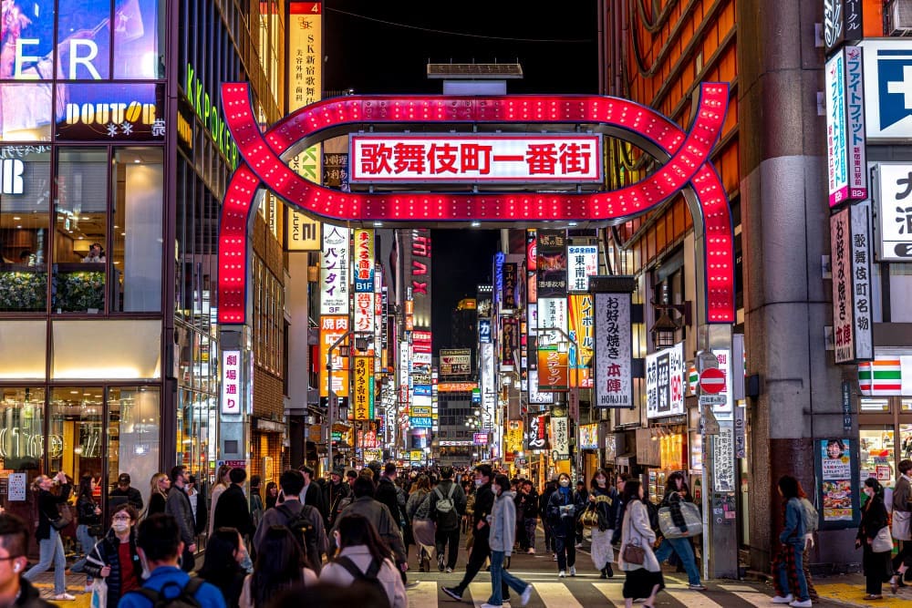 A vibrant nighttime street scene in Tokyo's Kabukicho district, with a large, illuminated red archway spanning the street, which is bustling with pedestrians and countless glowing neon signs.