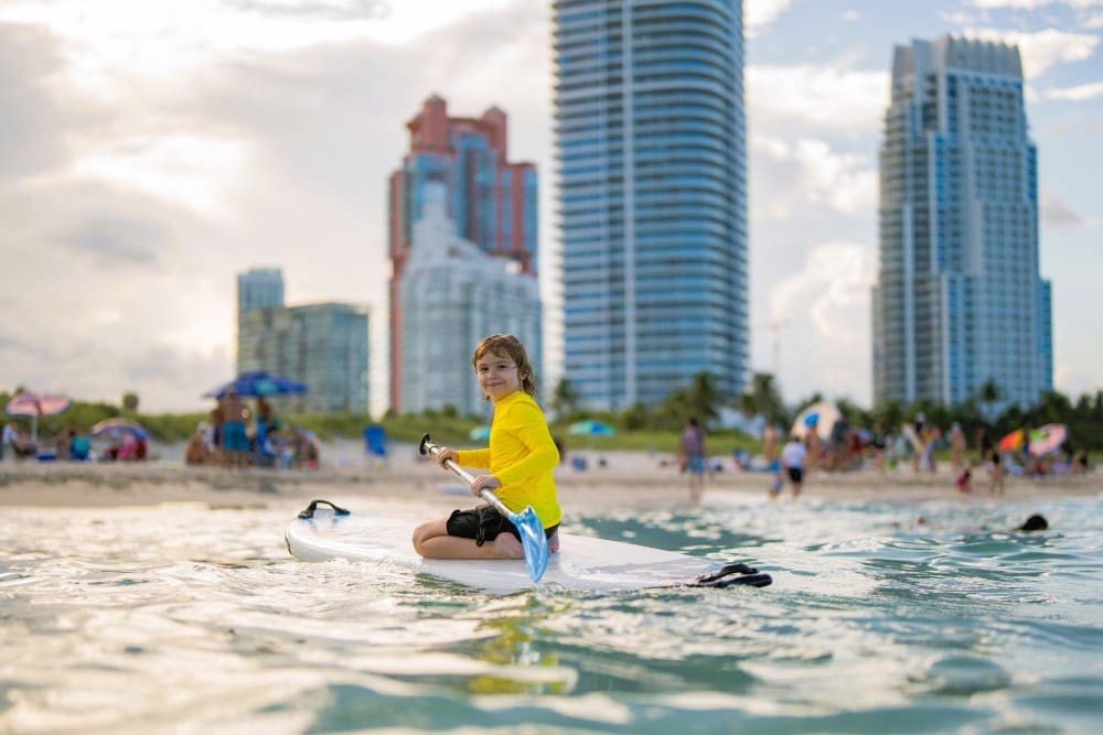 A smiling child in a yellow rash guard kneeling on a paddleboard in the ocean at South Pointe Beach, with buildings and beachgoers in the background.