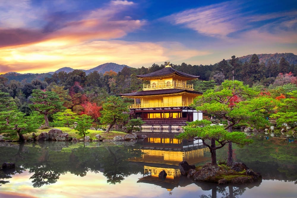 The shimmering golden Kinkaku-ji Temple (Golden Pavilion) stands by a calm pond, perfectly reflected in the water, surrounded by vibrant trees under a colorful sunset sky in Kyoto, Japan.