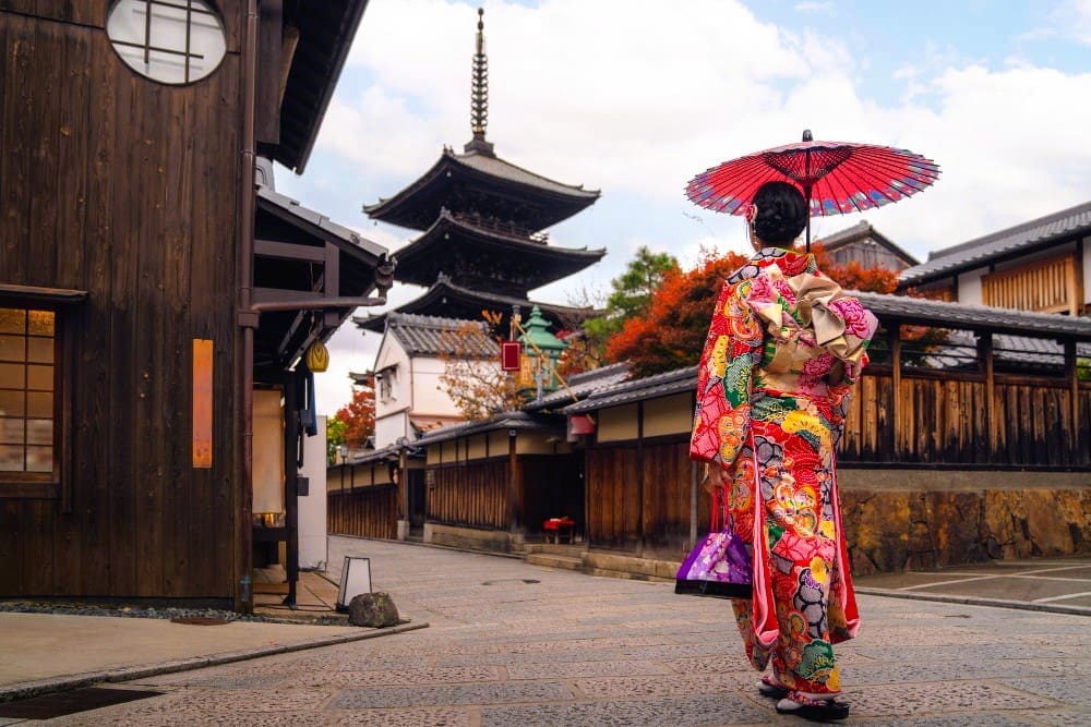 A woman in a vibrant kimono with a red parasol, walking towards a pagoda in a traditional Japanese town.
