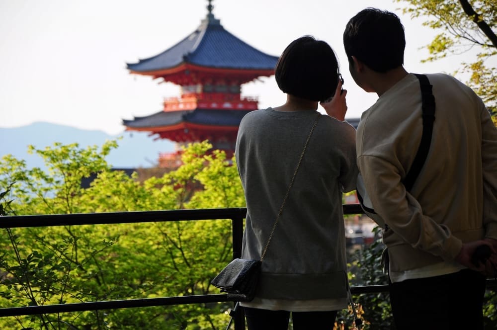 Rear view of a couple standing on a balcony, admiring a traditional red pagoda in Kyoto, Japan, surrounded by green foliage.