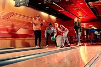 Four diverse young adults are enthusiastically bowling at a late-night alley with vibrant red and orange lighting, with one woman releasing a green bowling ball.