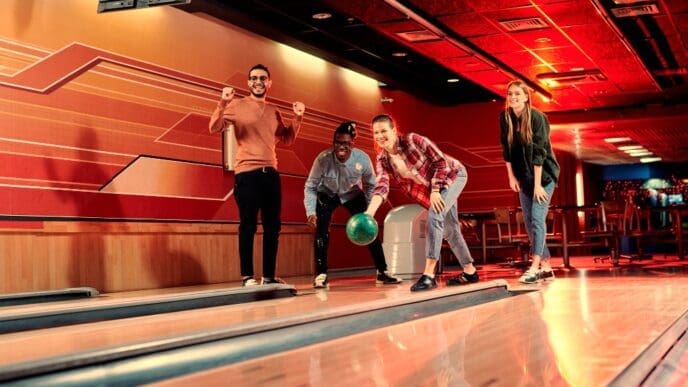 Four diverse young adults are enthusiastically bowling at a late-night alley with vibrant red and orange lighting, with one woman releasing a green bowling ball.