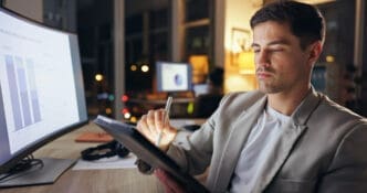 A focused man in a blazer working on a tablet with a stylus, looking at a computer monitor displaying graphs, in a dim office at night.