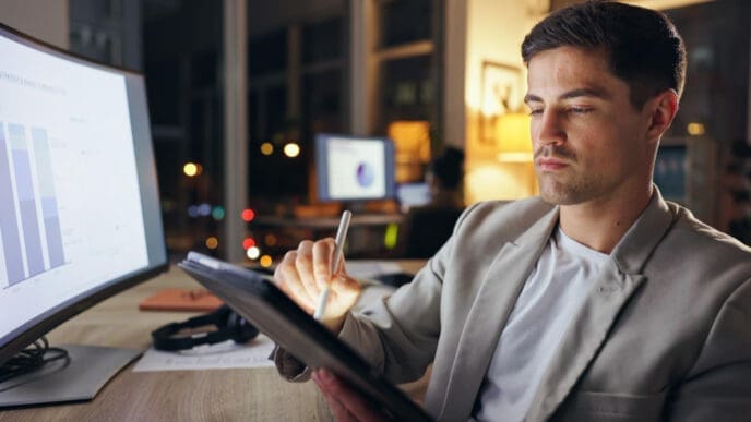 A focused man in a blazer working on a tablet with a stylus, looking at a computer monitor displaying graphs, in a dim office at night.