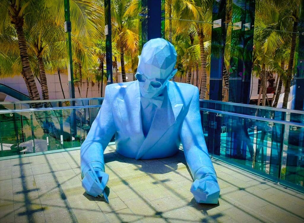 A modern, light blue geometric sculpture of a man in a suit with sunglasses, leaning on a table and writing, with lush palm trees and modern buildings in the vibrant sun-drenched background in Miami's Design District.